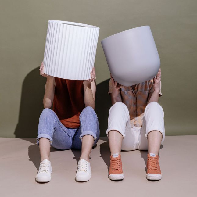 Two individuals with playful spirits sitting cross-legged, holding Slugg's unique white plant pots over their heads, showcasing the pots' sleek design and versatile style
